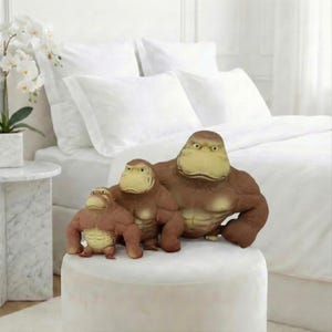 May include: Three brown gorilla figurines with tan faces and chests are displayed on a white ottoman. The gorillas vary in size, with the largest in the back and the smallest in the front. The background features a white bed and a marble side table.