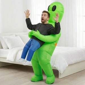 May include: An inflatable green alien costume with black eyes and a round orange detail on the side. The costume appears to be holding a person wearing a black shirt and blue jeans. The person has their arms raised and mouth open in surprise.