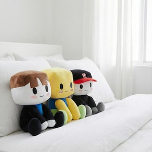 Könnte beinhalten: Drei Plüschpuppen mit quadratischen Köpfen und lächelnden Gesichtern sitzen auf einem weißen Bett. Die Puppen haben unterschiedliche Haarfarben, Kleidung und Accessoires, darunter eine rote Baseballkappe. Die Puppen sind schwarz, blau, gelb und braun.