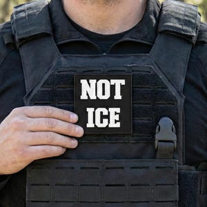 Könnte beinhalten: Schwarze taktische Weste mit dem Text "NOT ICE" in weißen Blockbuchstaben. Die Weste wird über einem schwarzen Hemd getragen. Der Hintergrund ist verschwommen, mit Grün- und Brauntönen.