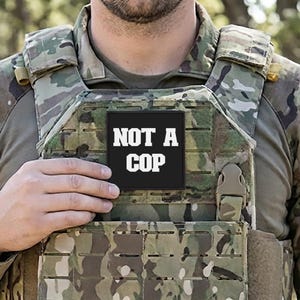 Puede incluir: Un parche cuadrado negro con las palabras "NOT A COP" en letras mayúsculas blancas en negrita. El parche está adherido a un chaleco táctico de camuflaje. La imagen es un primer plano, centrándose en el parche y el chaleco.