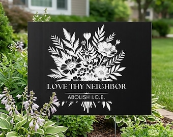 以下が含まれることがあります： 白い花の模様と「LOVE THY NEIGHBOR」と「ABOLISH I.C.E.」の文字が入った黒い庭の看板。看板は庭に設置されており、緑の葉や花に囲まれています。