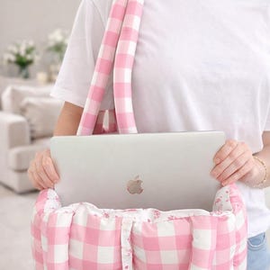 Może przedstawiać: R&oacute;żowa i biała torba typu tote w kratę z pasującymi, wyściełanymi paskami. Torba mieści srebrny laptop. Torba ma kwiatowe wykończenie i zapięcie na guziki. Torba jest przeznaczona do przenoszenia laptopa i innych niezbędnych rzeczy.