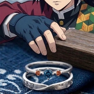 May include: Two silver rings with gemstones. One ring features a blue gemstone flanked by two orange gemstones. The other ring has a decorative design. The image also includes a character from an anime series.