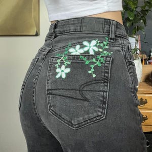 May include: Dark gray denim jeans with a floral embroidery design on the back pocket. The embroidery features white flowers and green vines, adding a touch of nature to the garment.
