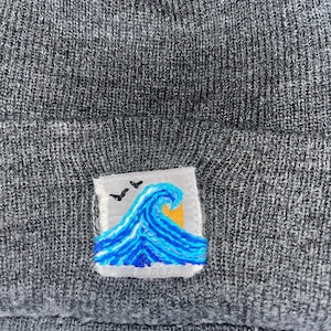 May include: Gray beanie with a small embroidered patch featuring a blue wave, yellow sun, and two black birds. The beanie is made of a knitted material.