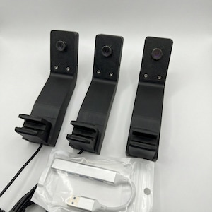 May include: Three black angled brackets with cameras, a silver USB hub, and black cables are shown against a white backdrop. The brackets have a distinctive design, with a camera at the top and a slotted base.
