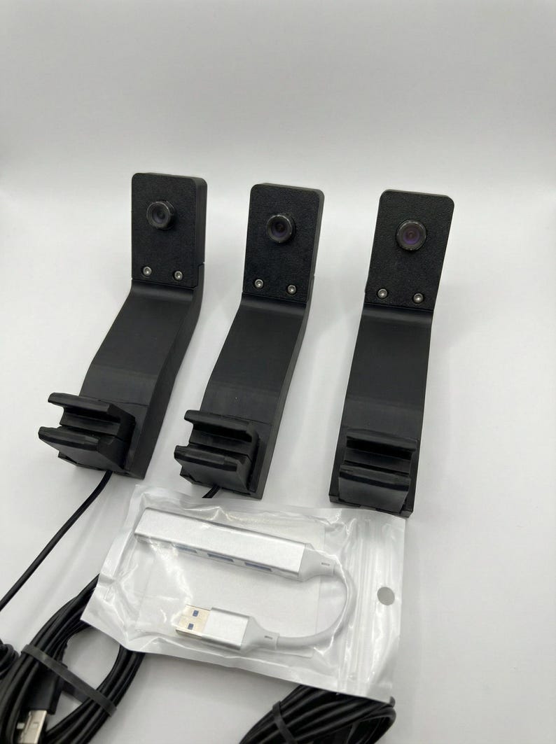 May include: Three black angled brackets with cameras, a silver USB hub, and black cables are shown against a white backdrop. The brackets have a distinctive design, with a camera at the top and a slotted base.