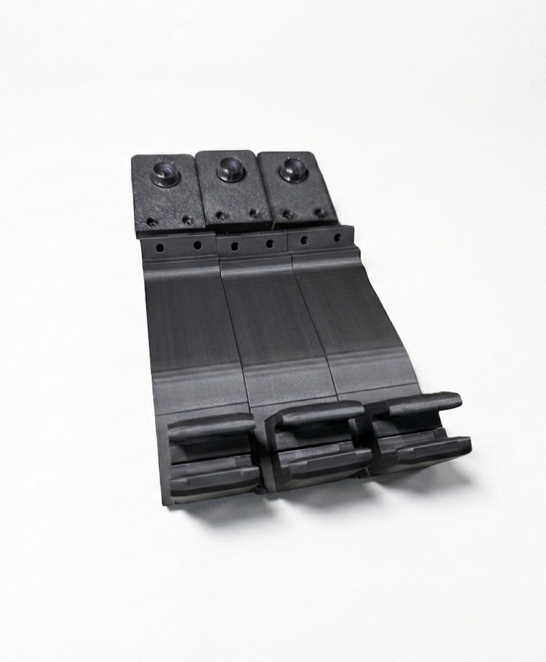 May include: A black, industrial component with three sections. Each section has a rectangular base with a curved top and small holes. The top features three raised circular elements. The component is set against a white background.