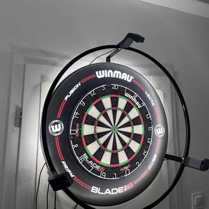 May include: A Winmau dartboard, featuring a black and red design with a green and white target. The board is mounted on a black metal frame with lights. The text "WINMAU" and "BLADE 6" are visible.