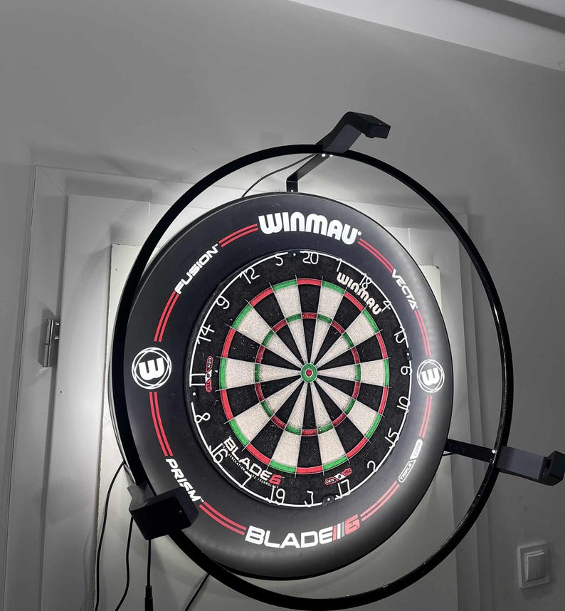 May include: A Winmau dartboard, featuring a black and red design with a green and white target. The board is mounted on a black metal frame with lights. The text "WINMAU" and "BLADE 6" are visible.