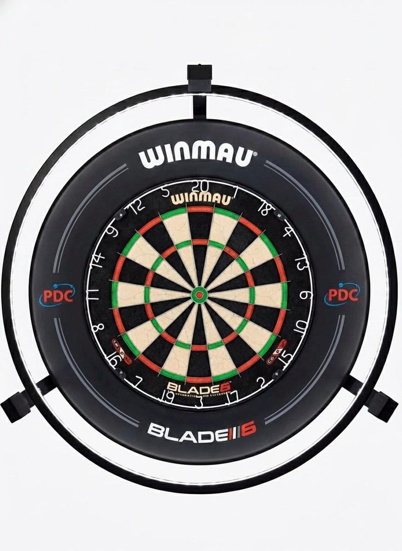 May include: A Winmau Blade 6 dartboard with a black surround and integrated lighting. The dartboard has a traditional design with red, green, and black segments. The Winmau logo is visible at the top of the board.