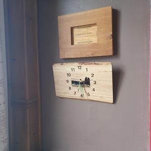 Op de afbeelding: Twee houten wanddecoraties. De bovenste is een rechthoekig frame met een vierkante opening. De onderste is een houten klok met zwarte cijfers en groene wijzers. De wijzerplaat heeft een kleine afbeelding van een gebouw.