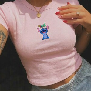 Peut inclure: Un crop top rose clair avec une image de dessin anim&eacute; de Stitch, un personnage extraterrestre bleu avec de grandes oreilles, tenant une petite plante. Le haut est &agrave; manches courtes et a une encolure ronde.