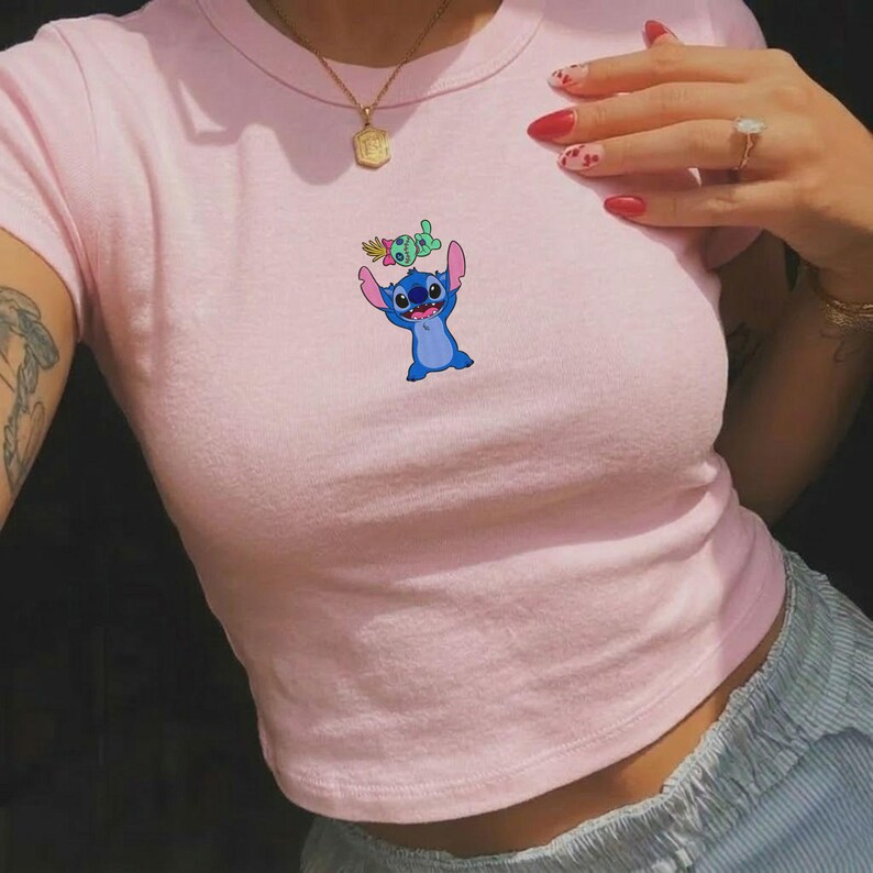 Peut inclure: Un crop top rose clair avec une image de dessin anim&eacute; de Stitch, un personnage extraterrestre bleu avec de grandes oreilles, tenant une petite plante. Le haut est &agrave; manches courtes et a une encolure ronde.