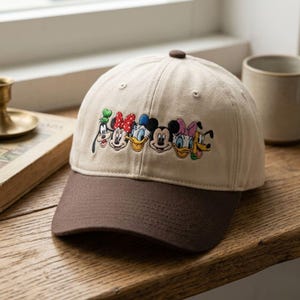 May include: A beige baseball cap with a dark brown brim featuring an embroidered row of six classic cartoon character faces including Goofy, Minnie Mouse, Donald Duck, Mickey Mouse, Daisy Duck, and Pluto across the front panel of the hat.