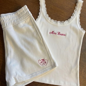 May include: A white ribbed tank top with lace trim and the embroidered text Mrs Evans in pink, paired with matching white shorts featuring an embroidered pink heart design with the letters A and H inside, creating a coordinated lounge set.