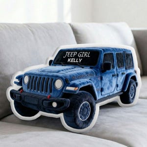 May include: A blue Jeep-shaped pillow with the text "JEEP GIRL KELLY" on the windshield. The pillow has a plush texture and features details like the Rubicon logo and headlights. It is resting on a light gray couch.