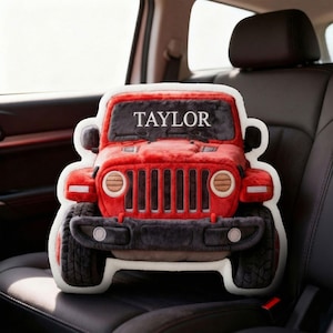 May include: A red Jeep-shaped plush pillow with the name "TAYLOR" across the top. The pillow has black and gray accents, including the tires and grill. The pillow is sitting in a car seat.