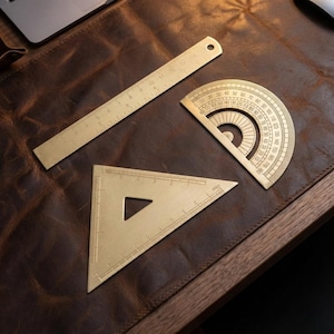 May include: A set of three brass drafting tools: a ruler, a protractor, and a triangle. The ruler is approximately 12 inches long. The protractor has degree markings, and the triangle has a cut-out. All tools are on a brown leather surface.