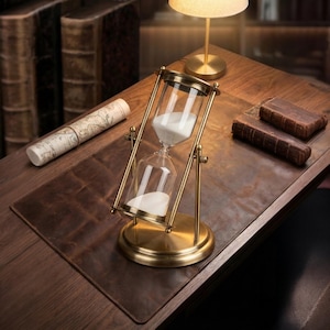 Può includere: Una clessidra in stile antico con struttura e base in ottone, riempita di sabbia bianca. Il timer in vetro è appoggiato su una scrivania in legno con un tappetino in pelle, accanto a una mappa arrotolata e due vecchi libri.