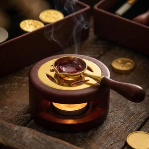 May include: A wooden wax warmer with a gold-colored metal plate and a small brass ladle filled with red wax. A lit tea light candle is inside the warmer, and smoke rises from the ladle. Gold wax seals are scattered around.