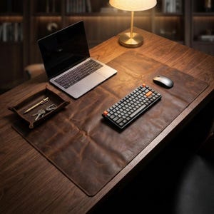 Puede incluir: Un tapete de escritorio de cuero marrón con una computadora portátil, un teclado, un ratón y una bandeja con un bolígrafo y llaves. El escritorio es de madera y tiene una pequeña lámpara. El teclado tiene teclas naranjas.