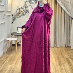 May include: A floor-length, long-sleeved magenta abaya featuring a subtle, repeating tonal jacquard pattern throughout the fabric. The garment includes a matching head covering that drapes over the shoulders and head, creating a cohesive look.