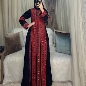 Puede incluir: Un vestido largo negro con intrincados bordados rojos. El vestido presenta patrones detallados en el corpiño, las mangas y la parte delantera. Las mangas y el escote están acentuados con bordados rojos. El vestido es largo hasta el suelo y parece estar hecho de una tela fluida.