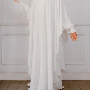 Puede incluir: Una abaya blanca hasta el suelo con una silueta holgada y fluida, con mangas anchas y drapeadas. La prenda tiene un cuello alto y puños fruncidos con volantes en las muñecas, creando una apariencia voluminosa en tela blanca sólida.