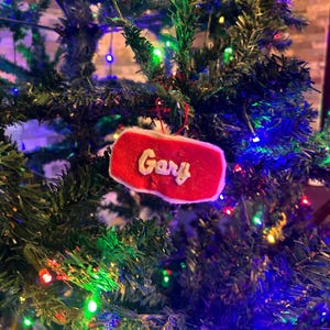 May include: A red Christmas ornament with the name "Gary" in white script. The ornament is rectangular with a white border and is hanging on a Christmas tree with colorful lights.