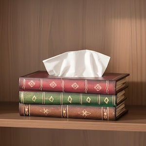 May include: A tissue box designed as a stack of old books. The box is made of wood and has a red, green, and brown colour scheme with gold detailing. White tissues are visible at the top, ready for use. The box sits on a wooden shelf.