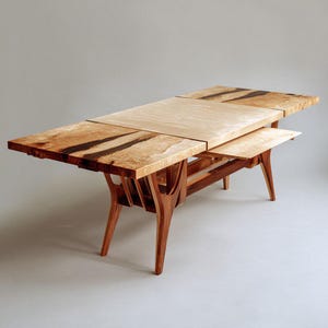 Może przedstawiać: Ręcznie wykonany drewniany stół jadalniany o unikalnym designie. Blat stołu posiada jasną, drewnianą część środkową, otoczoną ciemniejszymi, drewnianymi sekcjami z czarnymi akcentami. Stół ma charakterystyczną konstrukcję nóg i rozkładany blat. Stół ma około 183 cm długości.