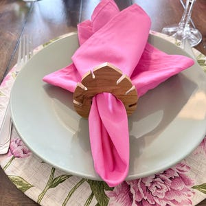 May include: A bright pink napkin is folded and held by a wooden leaf-shaped napkin ring. The napkin sits on a light green plate, atop a floral placemat with pink peonies. A fork and wine glass are visible.
