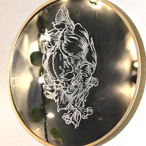 May include: Round mirror with a gold-colored frame. The mirror's surface is dark with a white, detailed floral and skull design etched into it. The design is centered on the mirror. The mirror is hanging on a white wall.