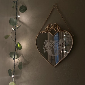 以下が含まれることがあります： 金色のフレームが付いたハート型の鏡が壁に掛けられています。鏡の表面には、翼のある2羽の白鳥のデザインが映っています。鏡の左側には、ライトのストリングと人工的な緑があります。