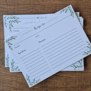 Puede incluir: Una pila de tarjetas de recetas blancas con diseños florales verdes en las esquinas. Las tarjetas están impresas con líneas para ingredientes, instrucciones y tiempo de preparación. Las palabras "Recipe for" están impresas en la parte superior de la tarjeta.