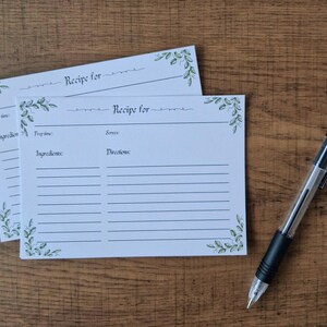 May include: Two white recipe cards with green floral accents and the text "Recipe for". A black and clear pen rests on a wooden surface. The cards have spaces for ingredients and directions.
