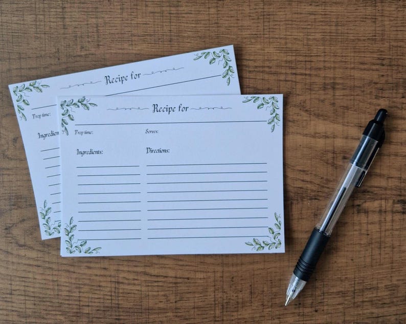 May include: Two white recipe cards with green floral accents and the text "Recipe for". A black and clear pen rests on a wooden surface. The cards have spaces for ingredients and directions.