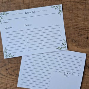 May include: Two white recipe cards with spaces for ingredients and directions. The top card reads "Recipe for" and has green leaf accents. The bottom card has a space for notes. Both cards are on a wooden surface.