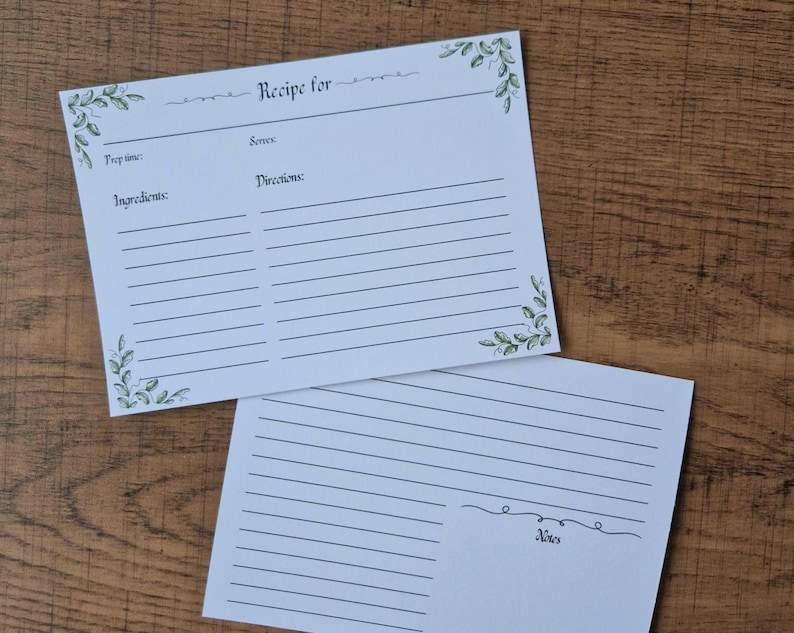 May include: Two white recipe cards with spaces for ingredients and directions. The top card reads "Recipe for" and has green leaf accents. The bottom card has a space for notes. Both cards are on a wooden surface.