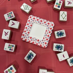 May include: A collection of white Mahjong tiles with various colorful symbols and numbers, scattered on a red surface. A decorative frame with a scalloped edge and a heart and star pattern is in the center. The tiles feature green, blue, and red accents.