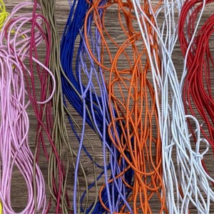 May include: A selection of colourful elastic cords in shades of yellow, pink, blue, orange, white, red, and black. The cords are arranged in a loose, tangled pattern against a wooden background, suggesting craft supplies.