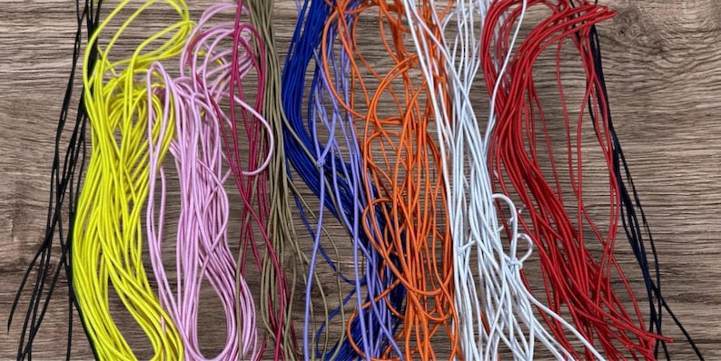 May include: A selection of colourful elastic cords in shades of yellow, pink, blue, orange, white, red, and black. The cords are arranged in a loose, tangled pattern against a wooden background, suggesting craft supplies.