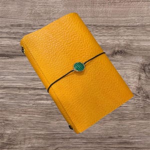 May include: A mustard yellow textured notebook cover secured with a thin black elastic cord. The cord features a circular charm with a green and black striped malachite-style inlay set in a gold-toned metal frame centered on the front cover.