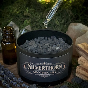 Puede incluir: Una lata cilíndrica negra llena de rocas volcánicas gris oscuro, con una etiqueta que dice Silverthorn Apothecary. Un gotero de vidrio dispensa un líquido transparente sobre la lata, junto a una pequeña botella de vidrio ámbar y lavanda.