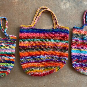 May include: Three handmade crochet tote bags of varying sizes, each featuring a vibrant, multi-colored horizontal striped pattern. The bags are constructed with a textured yarn weave, showcasing a spectrum of bright colors including red, orange, yellow, green, blue, and purple throughout the fabric.