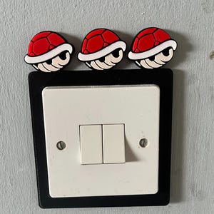 May include: A white double light switch mounted on a wall, framed by a black rectangular plastic cover. Three red and white turtle shell-shaped decorative toppers are attached to the top edge of the black frame, featuring a segmented shell design.
