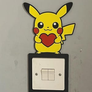 May include: A yellow Pikachu character cutout holding a red heart, positioned directly above a white double light switch with a black rectangular frame. The character features black-tipped ears, red circular cheeks, and a lightning bolt tail.
