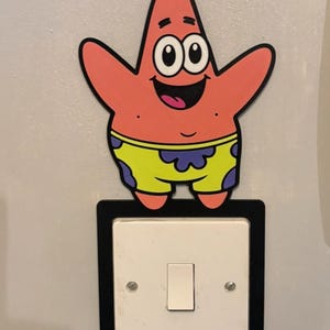 May include: A light switch surround featuring a cutout of the cartoon character Patrick Star. The character is pink with a smiling expression, wearing lime green shorts with a purple flower pattern. The surround has a black border around the white switch.